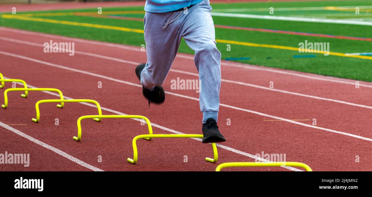 Vista frontale di un ragazzo della scuola superiore che corre su ostacoli gialli min su una pista rossa da vicino. Foto Stock