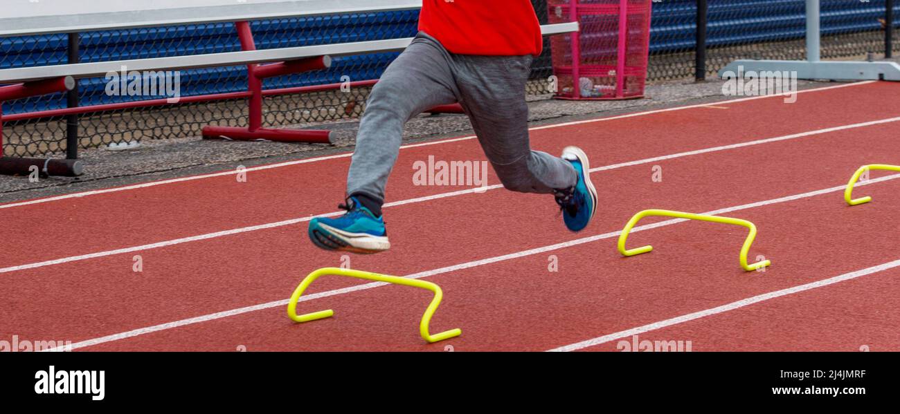 Un ragazzo di scuola superiore sta correndo sopra i piccoli ostacoli gialli mini su una pista. Foto Stock