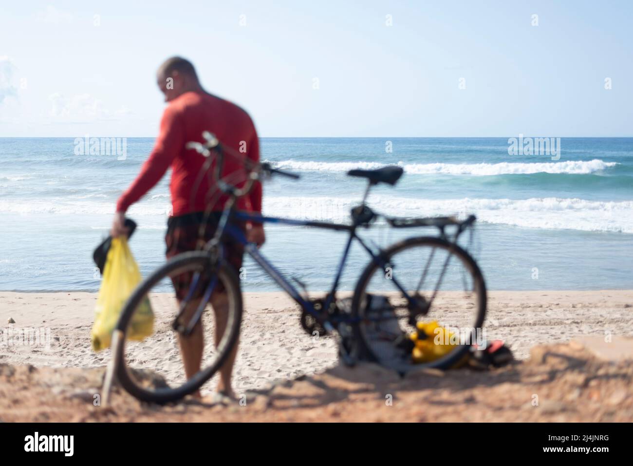 Un uomo in piedi sulla spiaggia accanto alla sua bicicletta con la spiaggia Boca do Rio sullo sfondo. Salvador, Bahia, Foto Stock