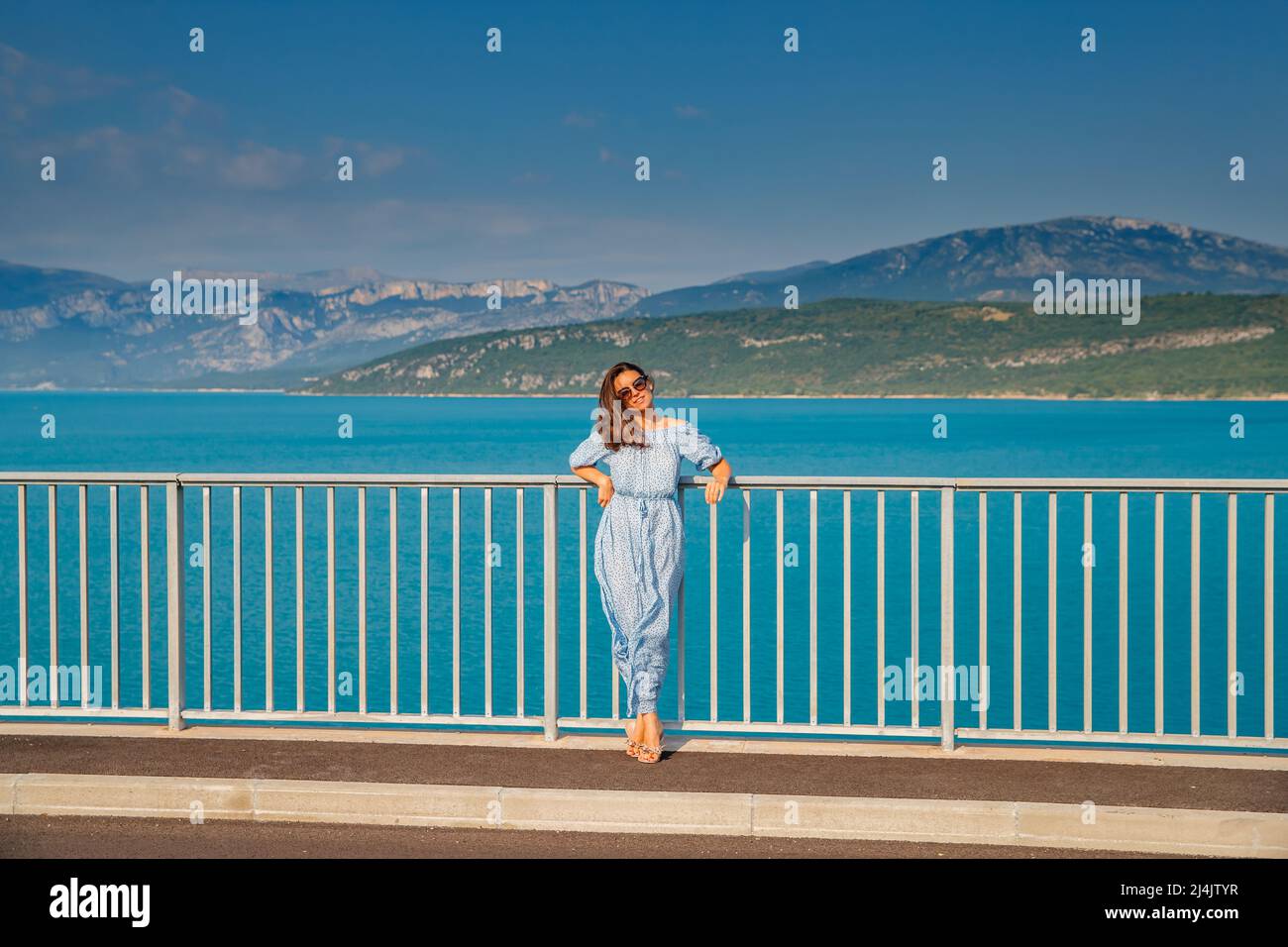 La bella ragazza in un abito blu e occhiali da sole pone sul ponte, i lunghi capelli castagno, felice e sorrisi, l'acqua azzurra del lago e pendii di Foto Stock