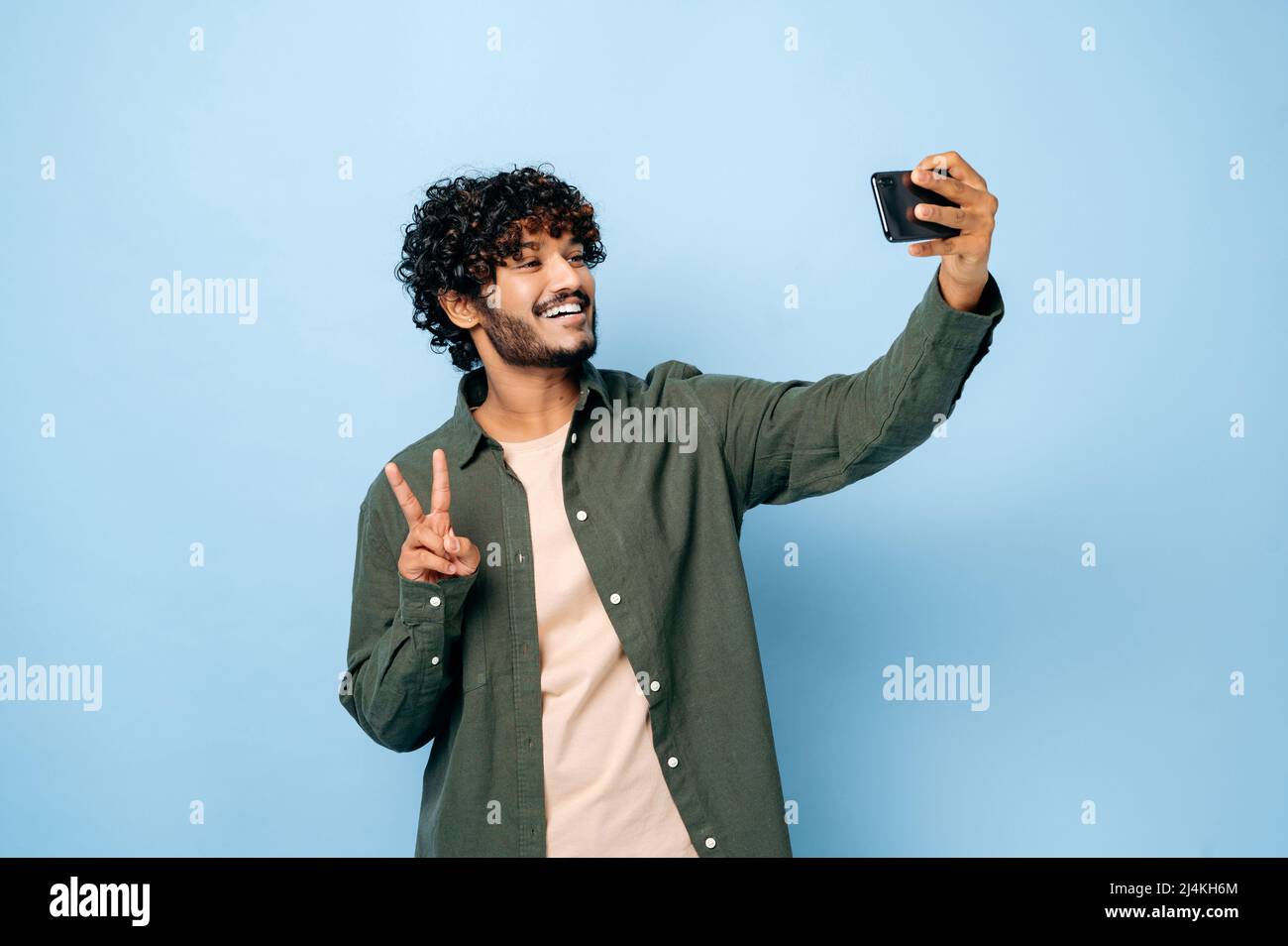 Allegro felice indiano o arabo eccitato ragazzo con gladden volto espressione, in abiti casual elegante, facendo selfie girato su smartphone e sorride nella fotocamera, si leva su isolato sfondo blu Foto Stock