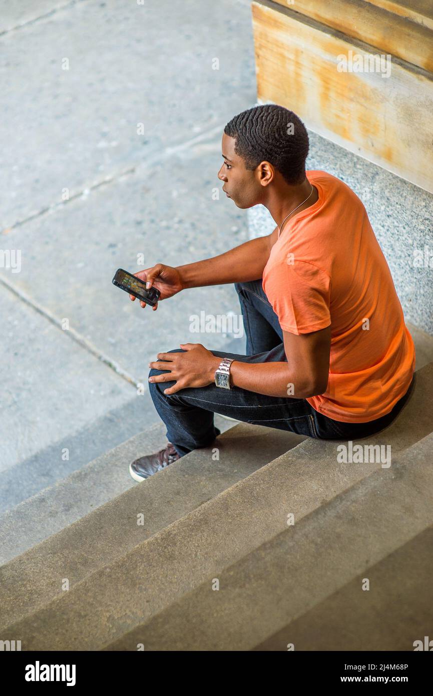 Un giovane e bello studente nero è seduto sulle scale all'angolo, controllando i messaggi sul suo cellulare e pensando Foto Stock