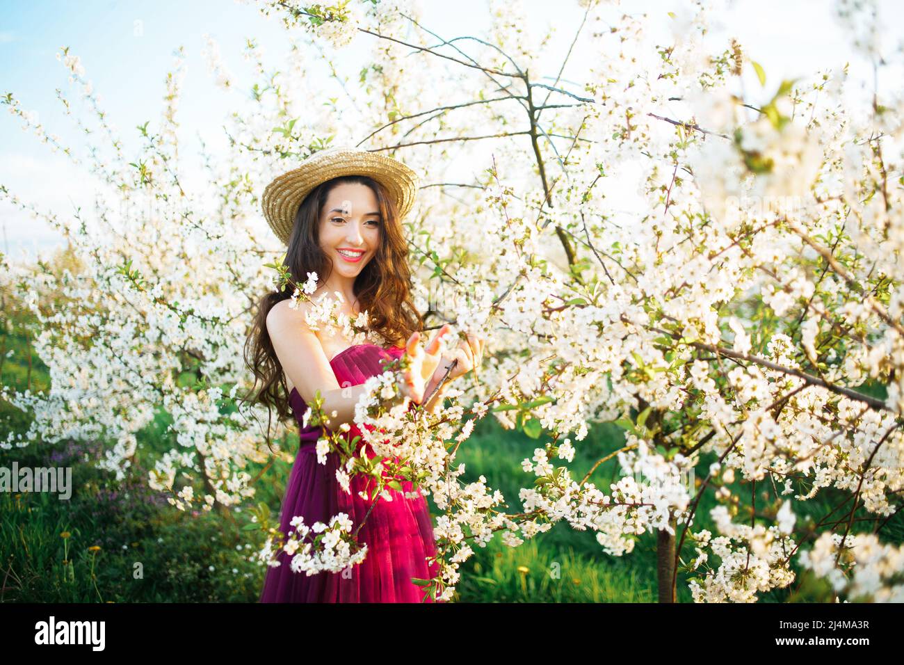 Ritratto di una ragazza sognante nel parco di primavera tra gli alberi bianchi fioriti al tramonto. Donna in cappello di paglia e abito viola, fiori di ciliegia. Molla s Foto Stock