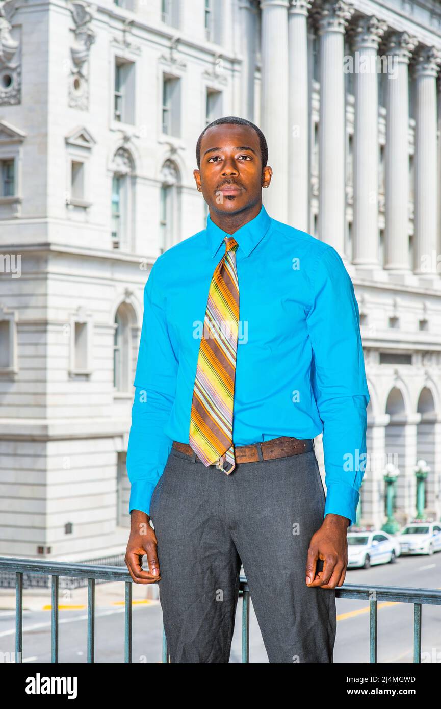 Vestendo in una camicia blu chiaro, pantaloni grigi, una cravatta colorata  motivo, un giovane uomo d'affari nero è in piedi fuori di un edificio  ufficio, guardando avanti Foto stock - Alamy