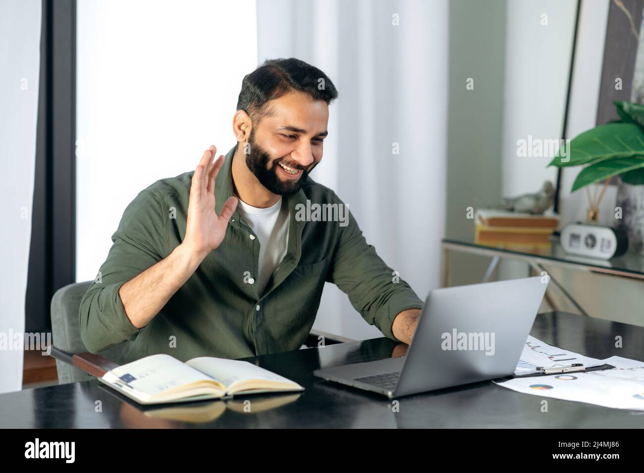 Simpatico ragazzo indiano amichevole, manager o consulente finanziario, comunica con i colleghi su una videochiamata, discute i risultati e la strategia in un ufficio moderno, negoziati a distanza, gesto di saluto Foto Stock