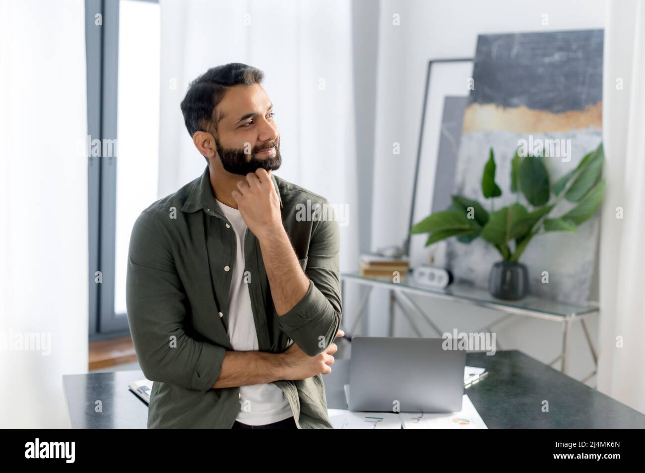 Ritratto di un uomo d'affari indiano o arabo di successo, manager o specialista IT, indossando abiti casual elegante, stand vicino alla sua scrivania in ufficio con le braccia incrociate, guardando lontano e sorridente Foto Stock