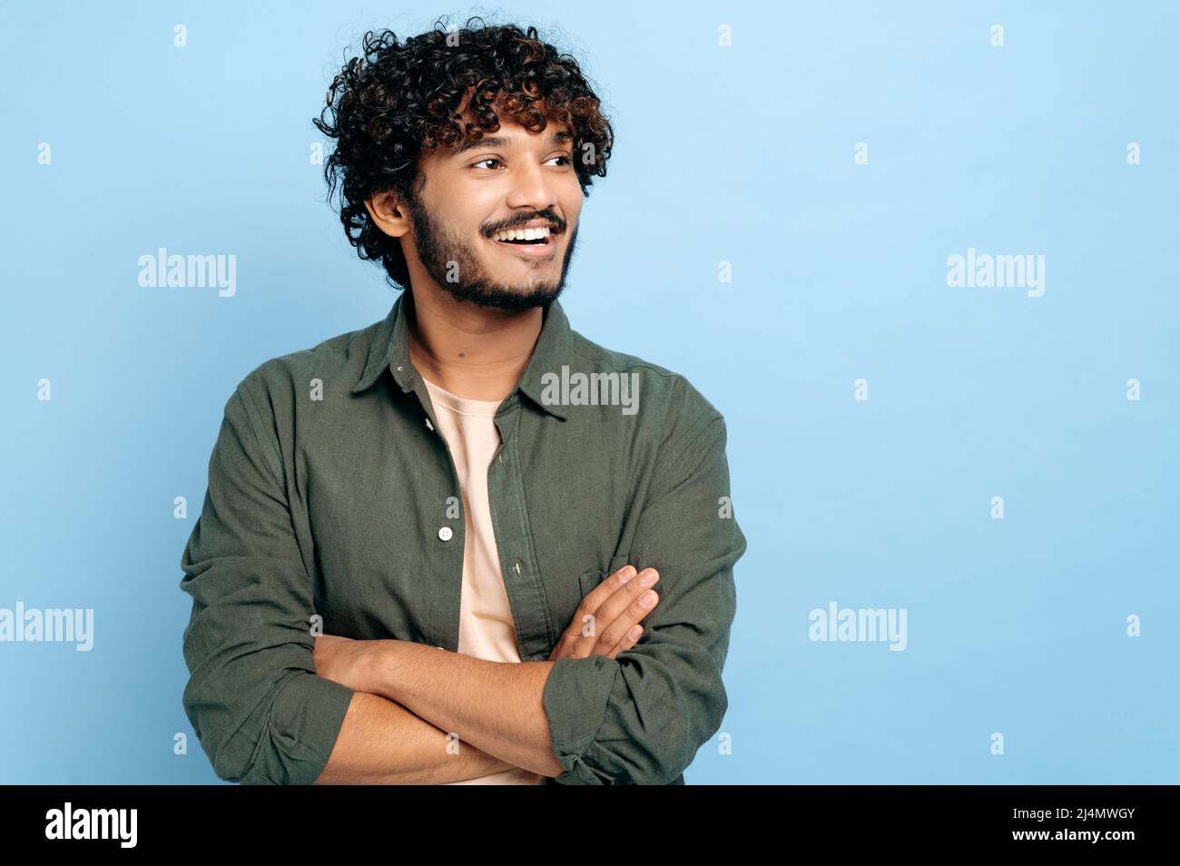 Bel ragazzo carismatico di successo indiano o arabo con capelli ricci in abiti eleganti, in piedi con le braccia incrociate su sfondo blu isolato, guardando via, sorridendo positivamente Foto Stock