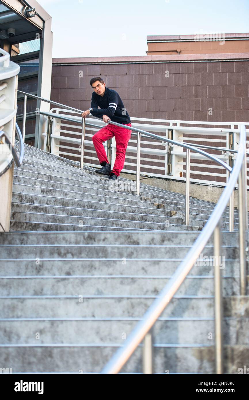 Vestendo in un maglione nero di colletto di scialle e jeans rossi, un giovane ragazzo bello è in piedi da ringhiere di una scala, in attesa di voi. Foto Stock