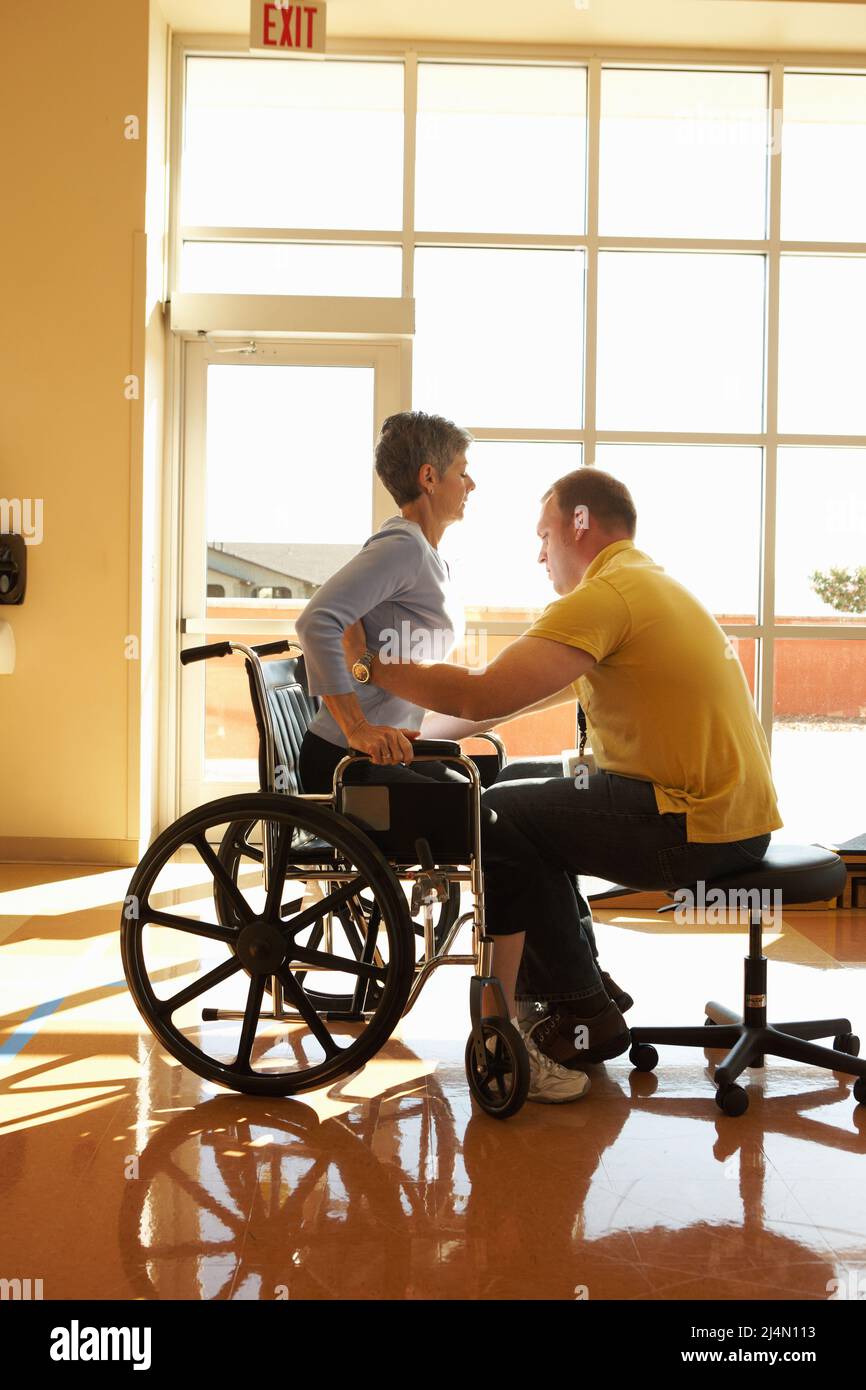 Fisioterapista che lavora con donna matura aiutandola ad imparare a alzarsi dalla sedia a rotelle in palestra ospedaliera progettata per riabilitare i pazienti che recuperano fr Foto Stock
