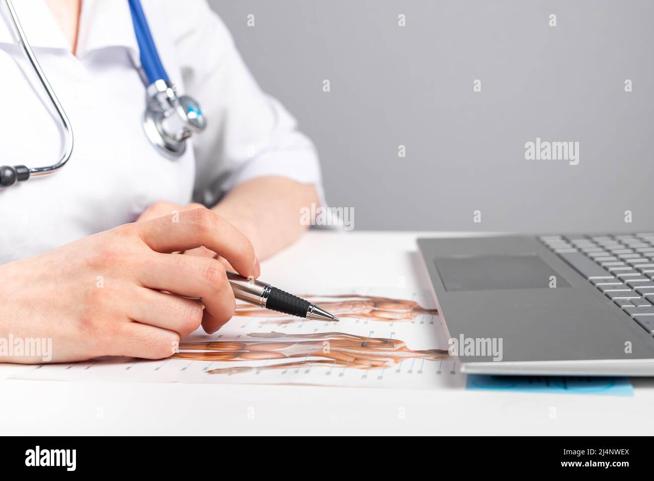 Dottore che studia o insegna anatomia. Educazione medica, concetto di corsi online. Donna in camice da laboratorio con stetoscopio seduto al tavolo con computer portatile e guardando carte con immagine del corpo umano. Foto Foto Stock