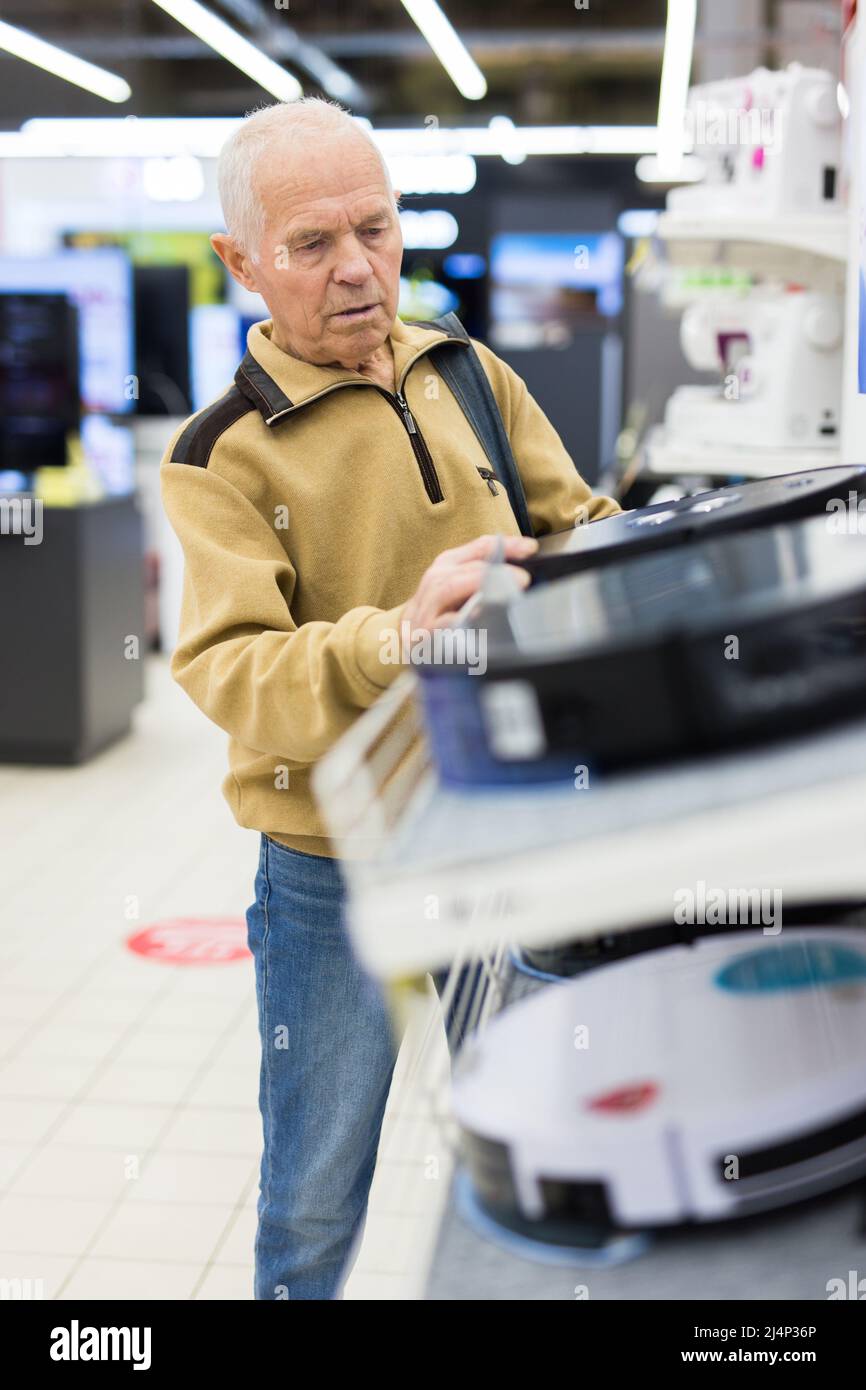 uomo anziano in grigio pensionato guardando robot hoover al banco in showroom di elettrodomestici ipermercato reparto Foto Stock