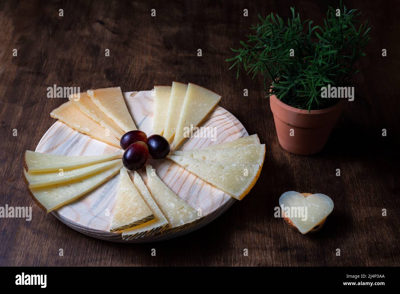 Bordo di formaggio spagnolo accanto ad una pianta in vaso, e un pezzo di formaggio a forma di cuore. Sfondo in legno. Foto Stock