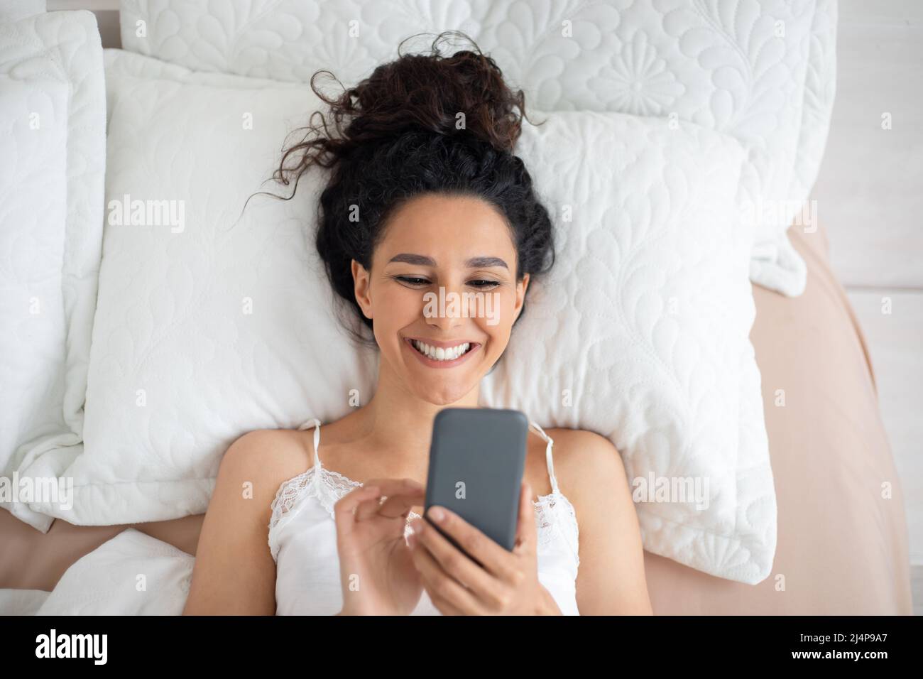 Allegra giovane donna bruna caucasica con capelli ricci in pigiama svegliarsi e leggere divertente messaggio sullo smartphone Foto Stock