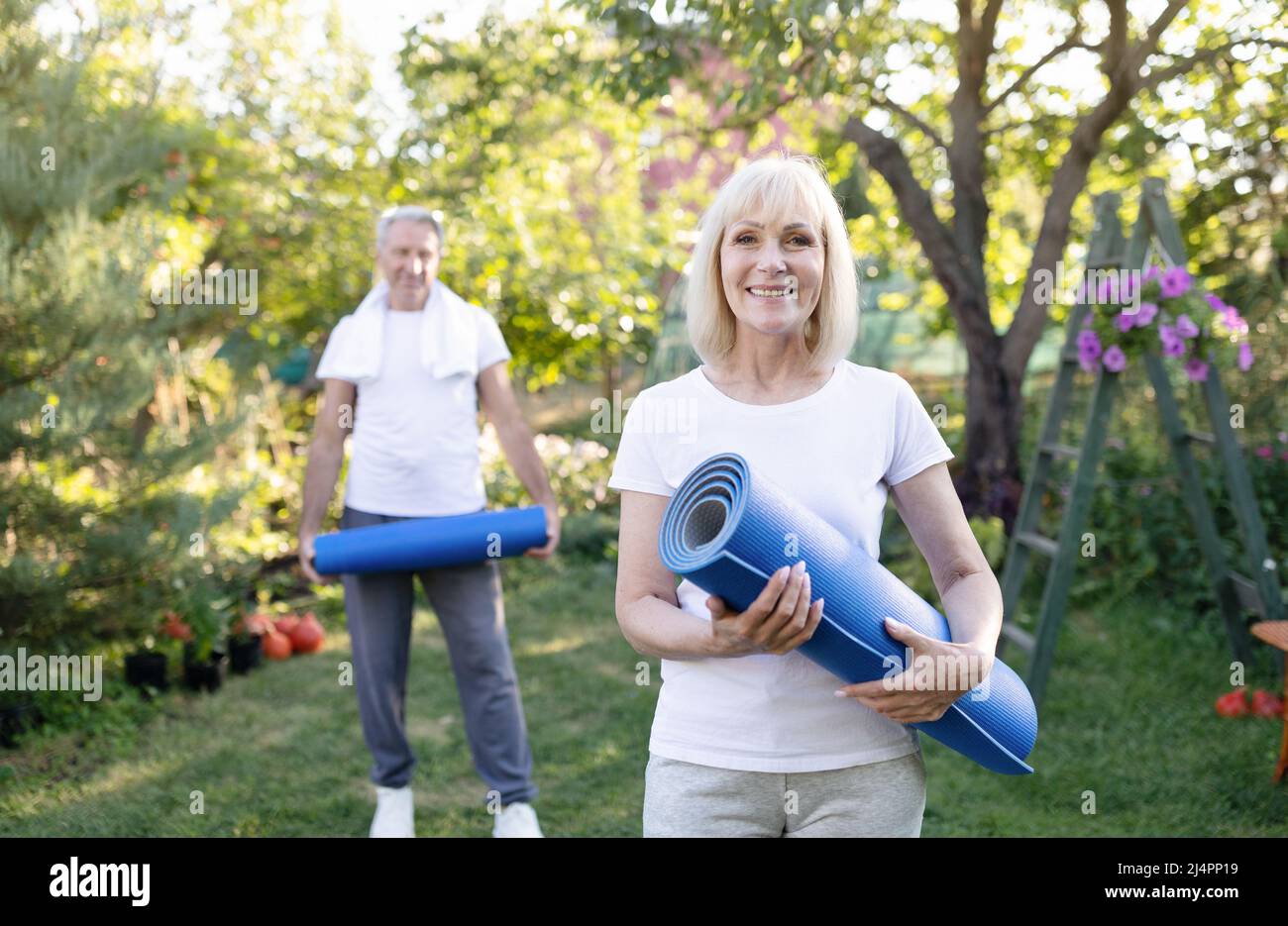 Ritratto di donna anziana attiva in posa con il marito in giardino, tenendo stuoie yoga, esercitandosi insieme all'esterno Foto Stock