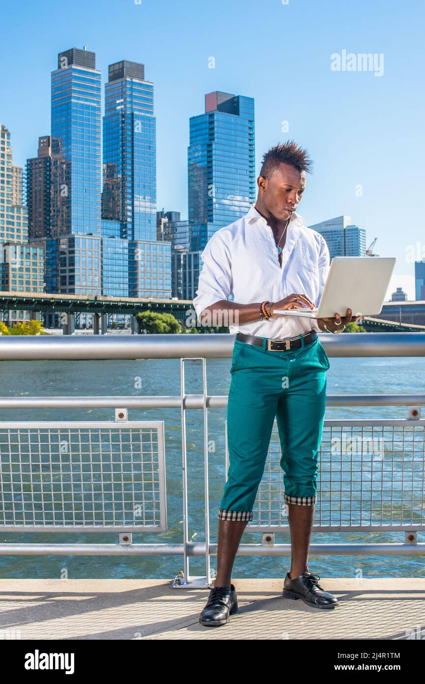 Vestendo in una camicia bianca, pantaloni verdi e scarpe di pelle, un giovane ragazzo nero con capelli mohawk è in piedi di fronte ad alti edifici, su cui si lavora Foto Stock