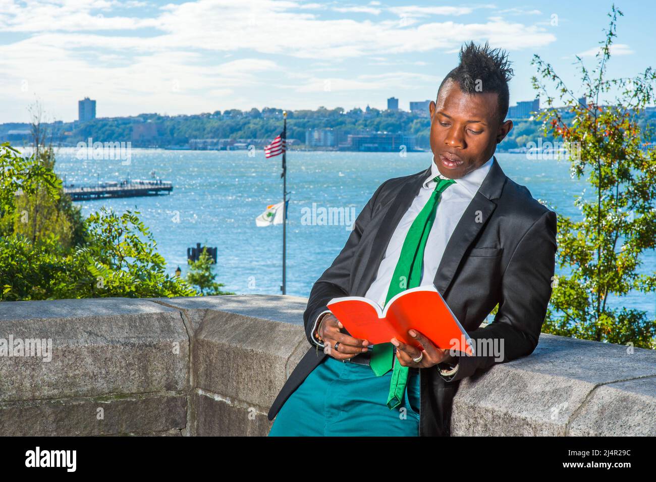 Il giovane uomo che legge fuori. Indossando un blazer nero, pantaloni verdi, una cravatta da collo verde, le mani che tengono un libro rosso, un giovane ragazzo nero con taglio di capelli mohawk è s Foto Stock