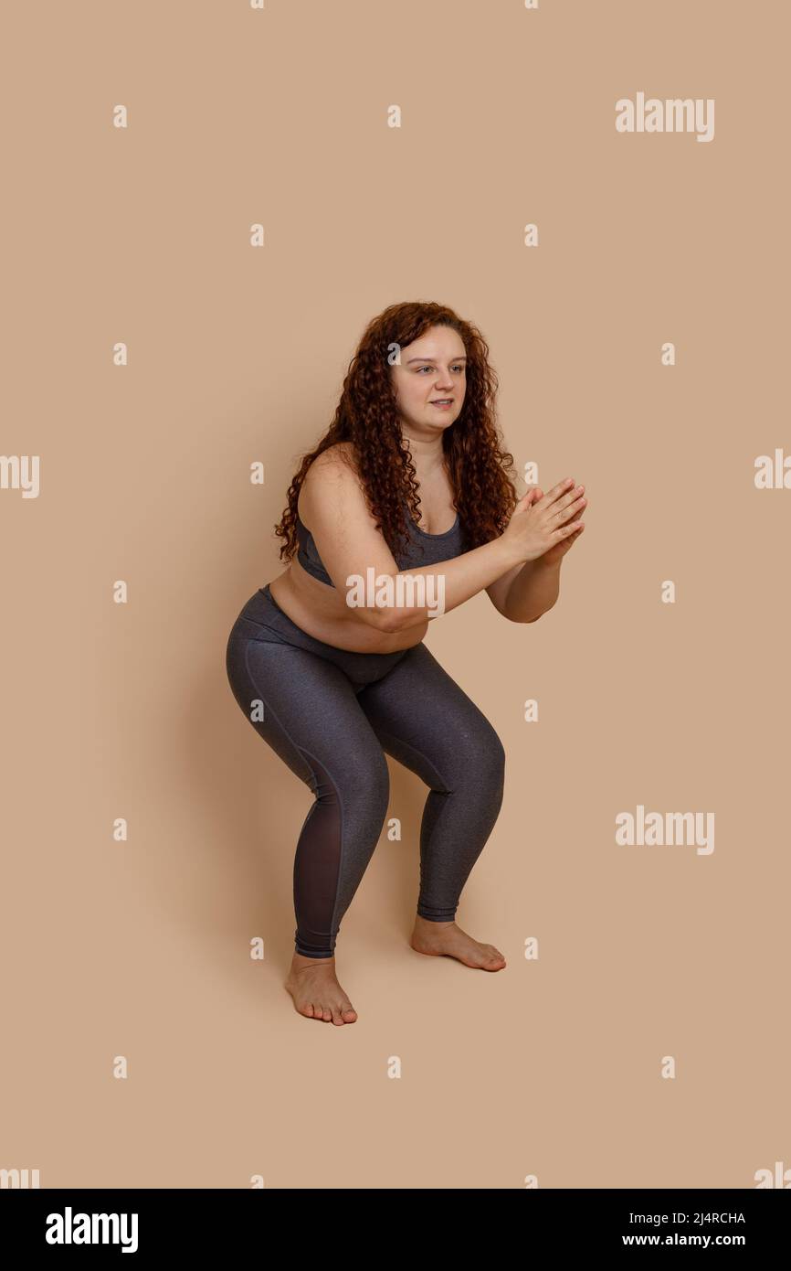 Immagine verticale di donna a piedi nudi focalizzata sui grassi, in gambali grigi e in cima. Piegamento del ginocchio, squat. Sport di salute positivo Foto Stock