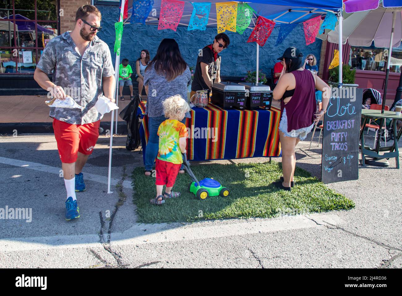 Bambino biondo pelato che rasa una toppa di erba artificiale di fronte a un cavalletto taco con un rasaerba giocattolo mentre gli adulti stanno comprando cibo a Cherry Street Farme Foto Stock
