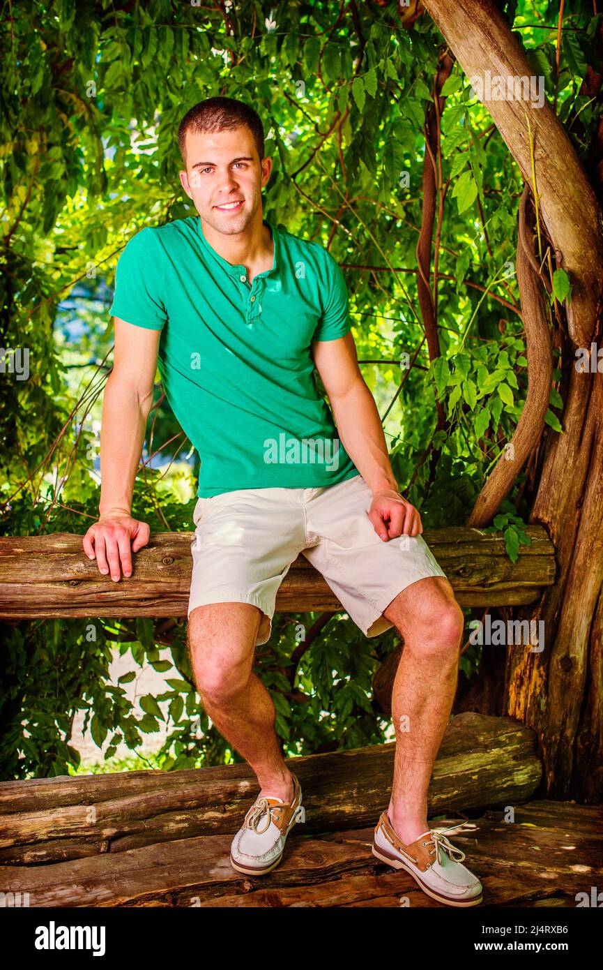 Giovane uomo che si rilassa all'esterno. Indossalo con camicia verde corta Henley, pantaloncini gialli chiari, scarpe casual in pelle, ragazzo giovane e bello seduto su tru albero Foto Stock
