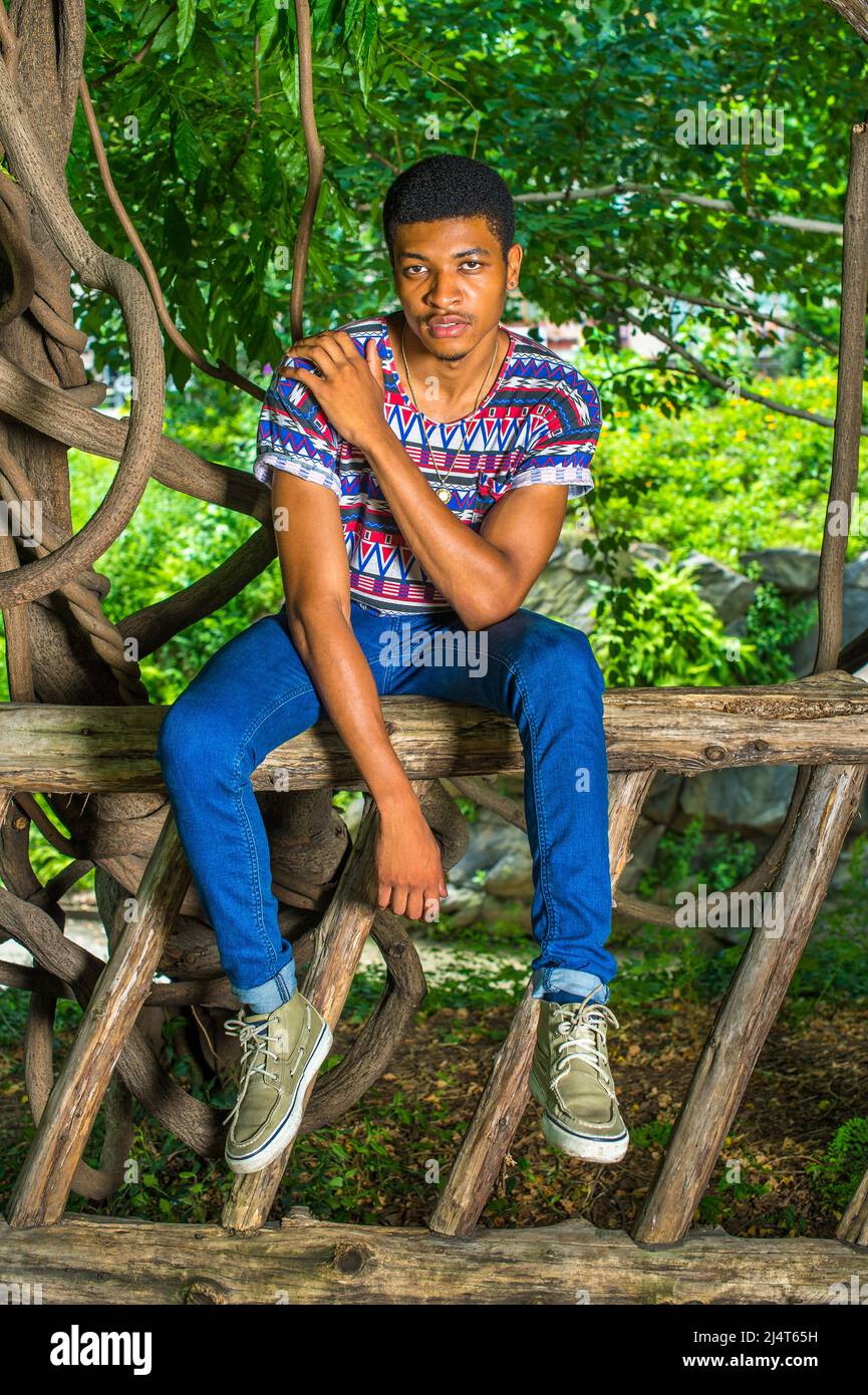 Black Man rilassante all'esterno. Indossando una camicia colorata, jeans blu, sneakers, collana, un giovane ragazzo bello è seduto su recinzione di legno, in attesa Foto Stock