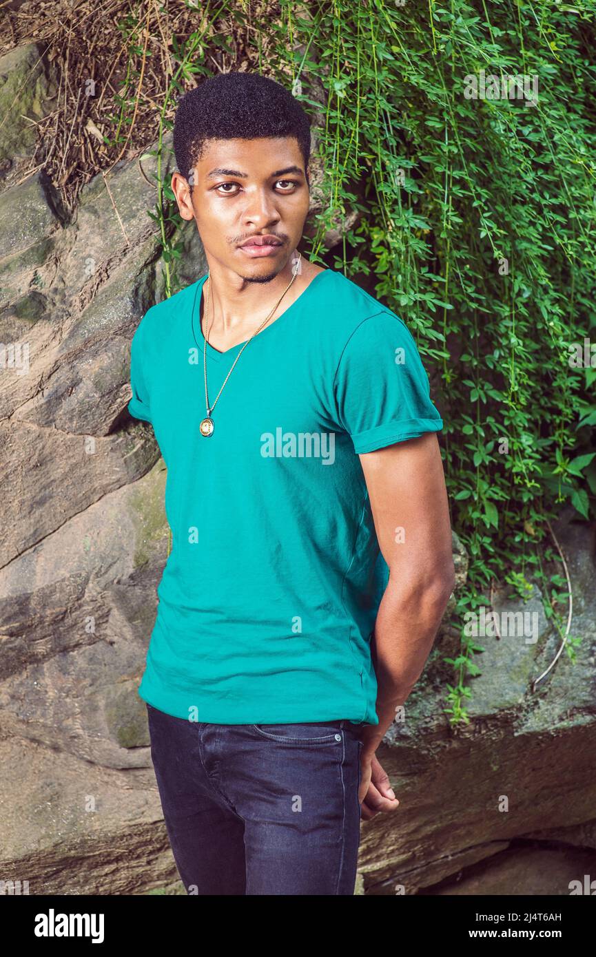 Ritratto di giovane uomo Nero. Indossando una manica corta verde, una T-shirt con collo a V, una collana, pantaloni neri, un ragazzo giovane e bello è in piedi da rocce con gree Foto Stock