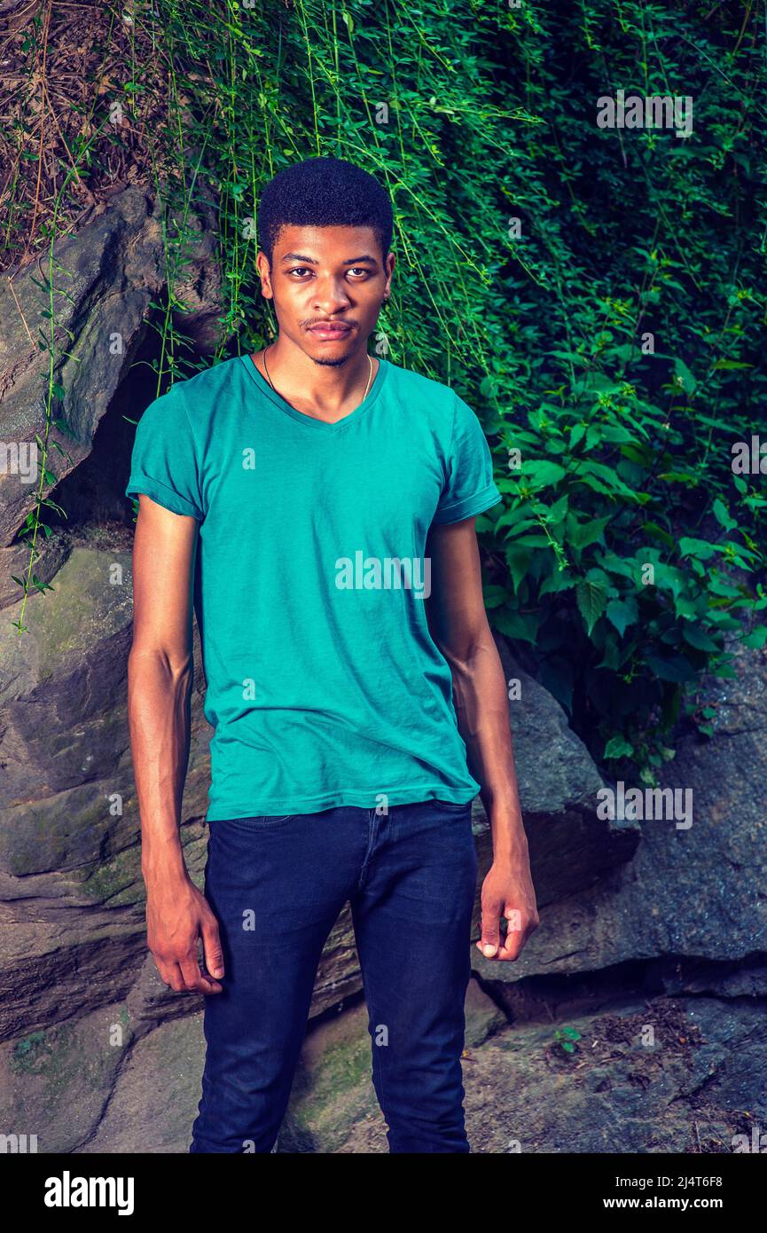 Ritratto di giovane uomo Nero. Indossando una manica corta verde, una T-shirt con collo a V, pantaloni neri, un giovane ragazzo bello è in piedi da rocce con foglie verdi, Foto Stock