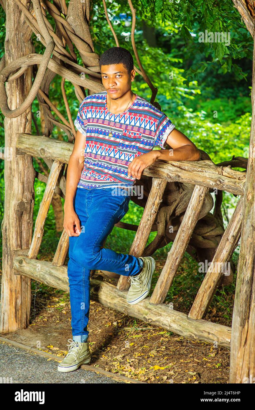 Giovane uomo Nero che ti aspetta. Indossando una camicia colorata modello, jeans blu, sneakers, collana, un giovane ragazzo bello è in piedi contro un f di legno Foto Stock