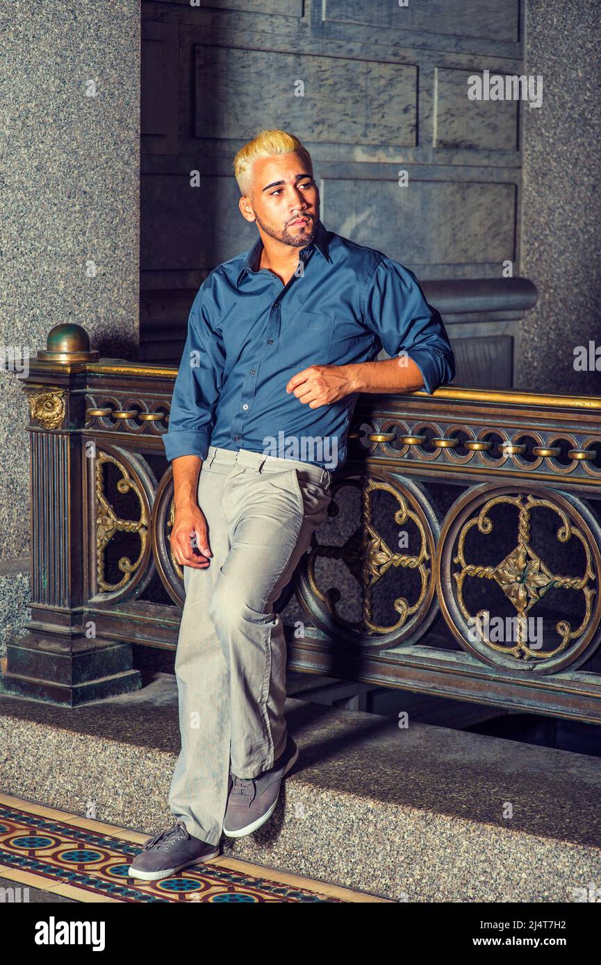 Uomo che ti aspetta. Indossando una camicia blu, pantaloni grigi, scarpe casual, un ragazzo giovane con barba, capelli gialli è in piedi da vecchio stile di moda ringhiera in un Foto Stock