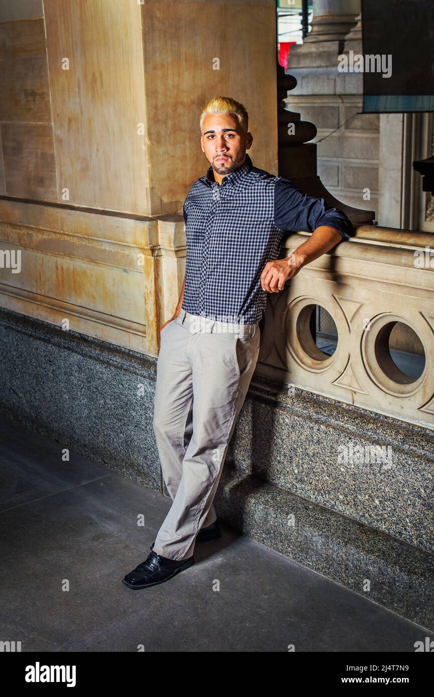 Ritratto di giovane lavoratore. Indossando una camicia a motivi neri, pantaloni grigi, scarpe in pelle, un ragazzo giovane con barba, capelli gialli, è in piedi da una ringhiera Foto Stock