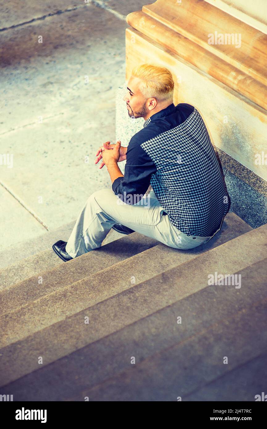 Giovane uomo che si rilassa all'esterno. Indossando una camicia a motivi neri, pantaloni grigi, scarpe in pelle, un ragazzo giovane con barba, capelli gialli, è seduto sulle scale, bac Foto Stock