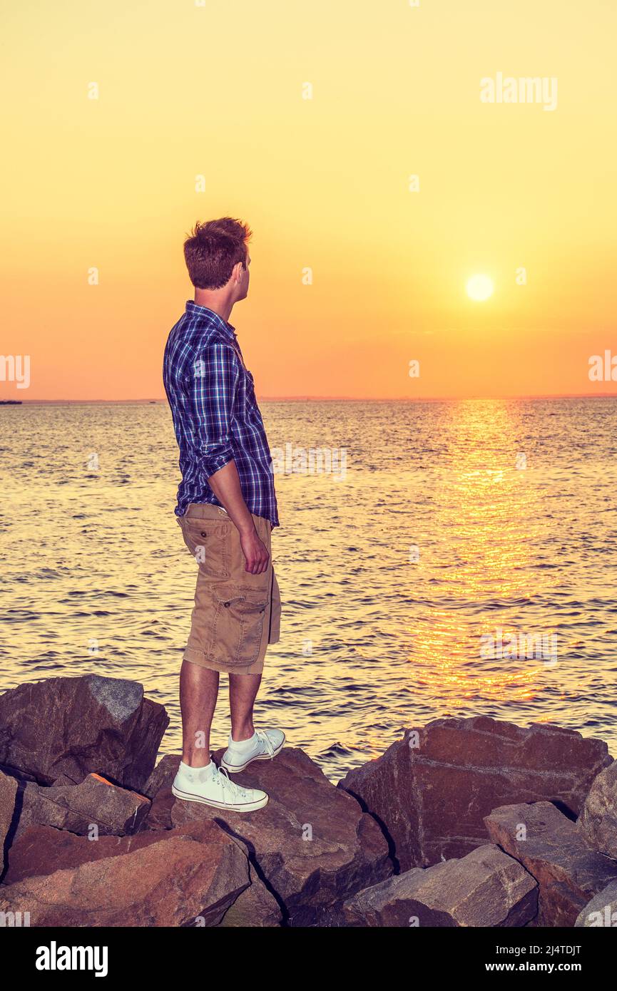 Uomo che guarda il tramonto nella vista posteriore. Indossando una camicia bianca, blu a motivi geometrici, pantaloni gialli, sneakers bianchi, un bel ragazzo è in piedi sulle rocce sulla spiaggia, loo Foto Stock