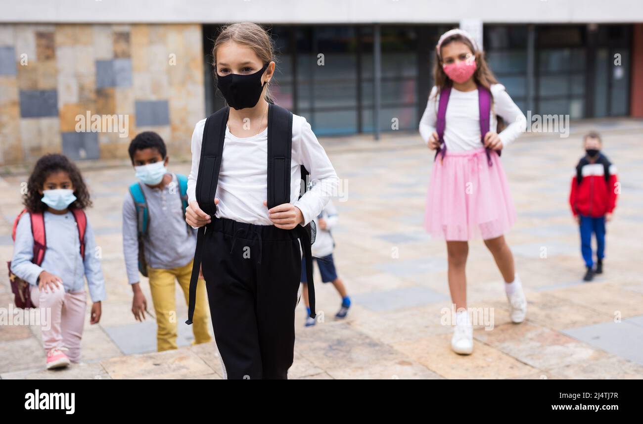 Studentessa in maschera medica in piedi sulla strada, bambini sullo sfondo Foto Stock