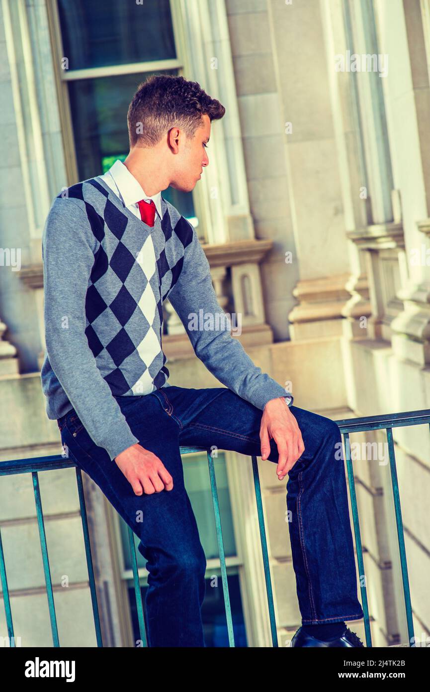 Uomo che si rilassa all'esterno. Vestendo in un nero, bianco, grigio, pullover a motivi geometrici, jeans, mani che riposano sui giri, un giovane uomo d'affari bello è seduto su un r Foto Stock