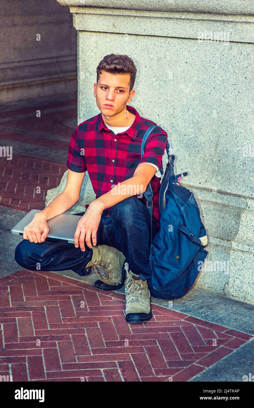 Vestirsi in manica corta, nera, camicia a motivi rossi, jeans, scarpe da calcio, trasportando la borsa della spalla, tenendo un calcolatore del laptop, un ragazzo giovane sta squatting Foto Stock