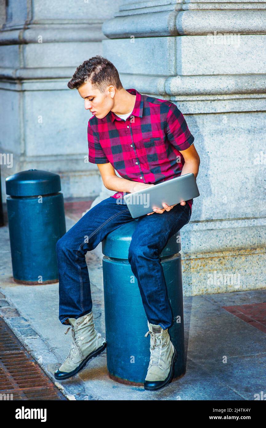 Distrazione. Indossando una manica corta, una camicia a motivi geometrici, jeans, scarpe da stivale, tenendo un computer portatile, un ragazzo è seduto su una colonna di metallo, guardando Foto Stock