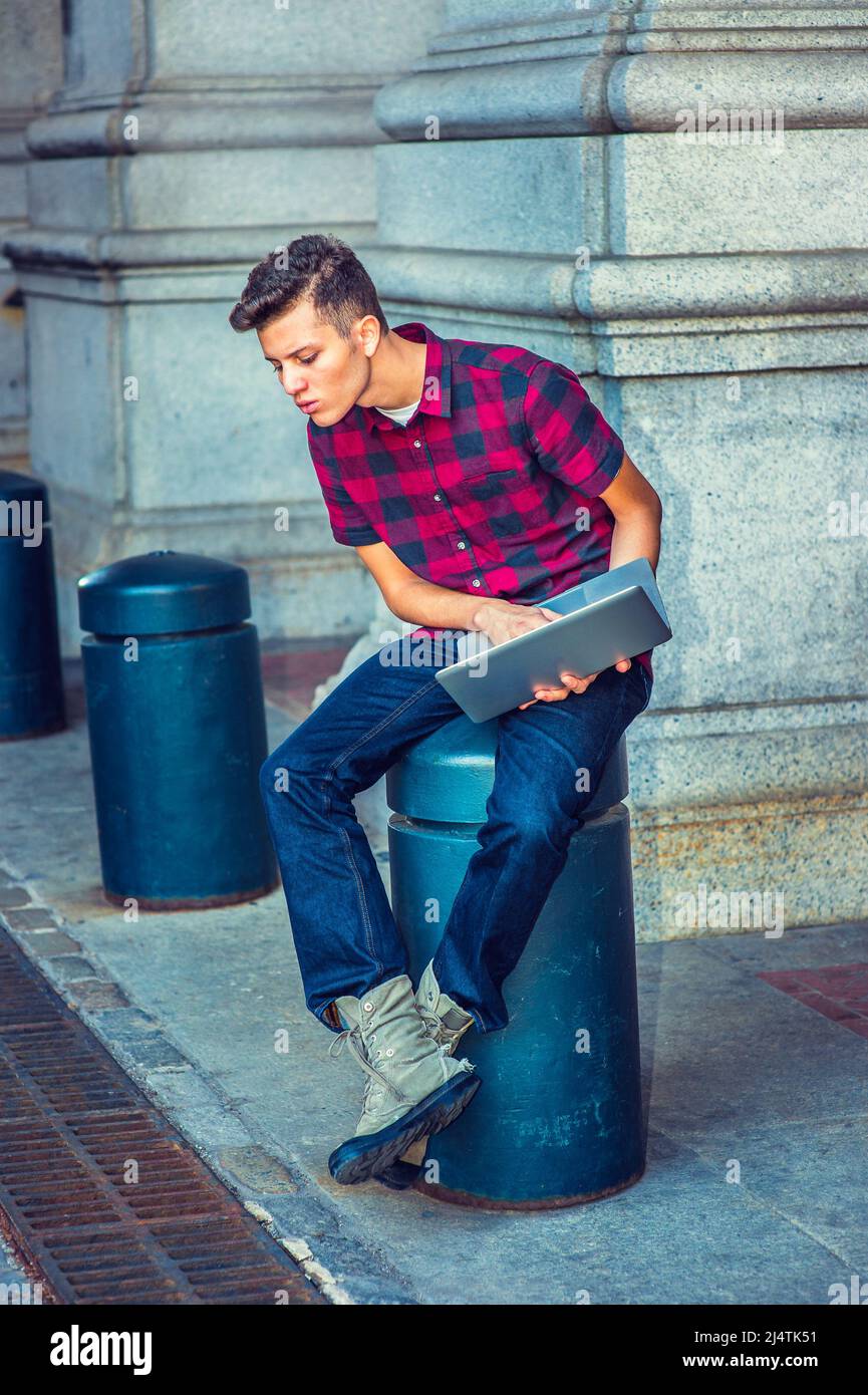 Distrazione. Indossando una manica corta, una camicia a motivi geometrici, jeans, scarpe da stivale, tenendo un computer portatile, un ragazzo è seduto su una colonna di metallo, guardando Foto Stock