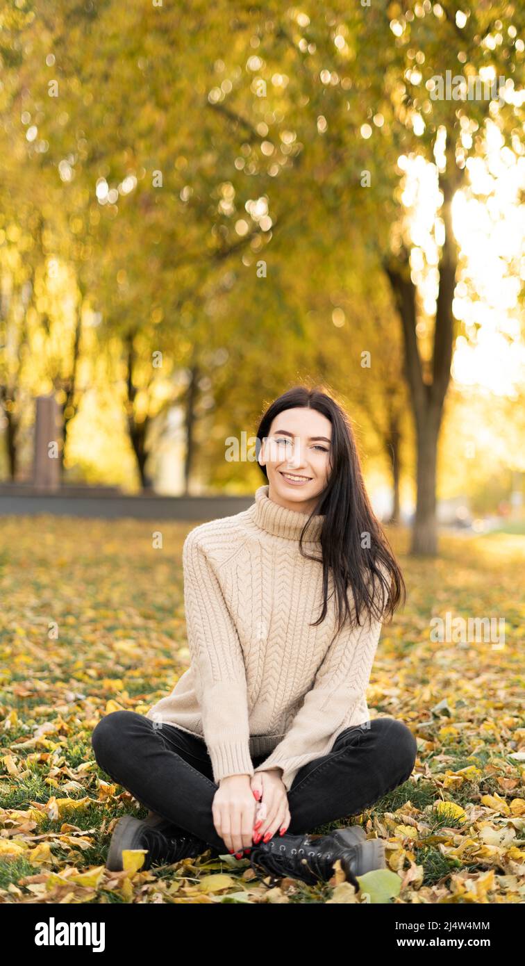 Una ragazza giovane e bella con capelli lunghi siede in molte foglie di arancio e giallo caduto a terra in un parco. La donna indossa un maglione. Gioia, autum Foto Stock