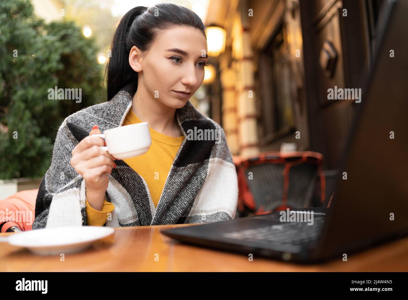 Una ragazza in un bar sulla strada avvolto in una plaid, godendo di una calda giornata di sole autunno. Tazza di caffè in mani giovane brunetta che lavora dietro un computer portatile Foto Stock