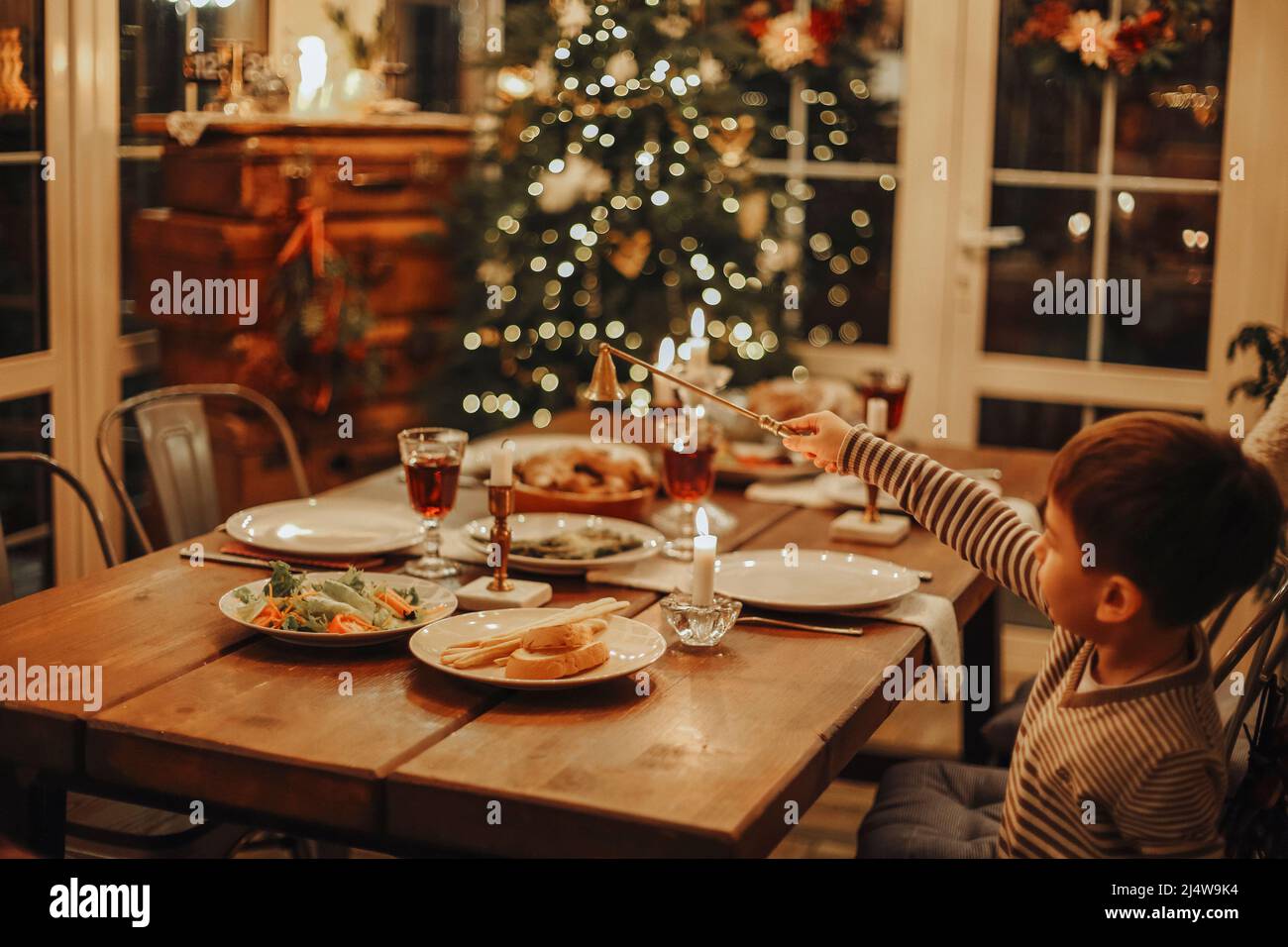 Il ragazzo piccolo divertente è seduto sul tavolo della cucina di Natale in un maglione bianco che mette fuori la fiamma della candela con campanello snuffer dopo la celebrazione del Natale h Foto Stock