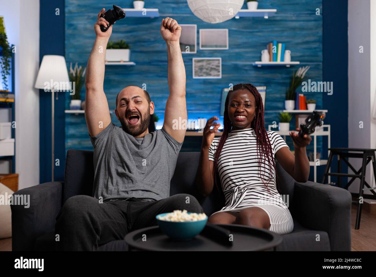 Uomo caucasico felice per vincere gioco di console contro la ragazza afro-americana mentre si siede a casa sul divano. Eccitata maschio adulto sopra la vittoria del video gioco mentre donna sconvolto a causa di perdere. Foto Stock