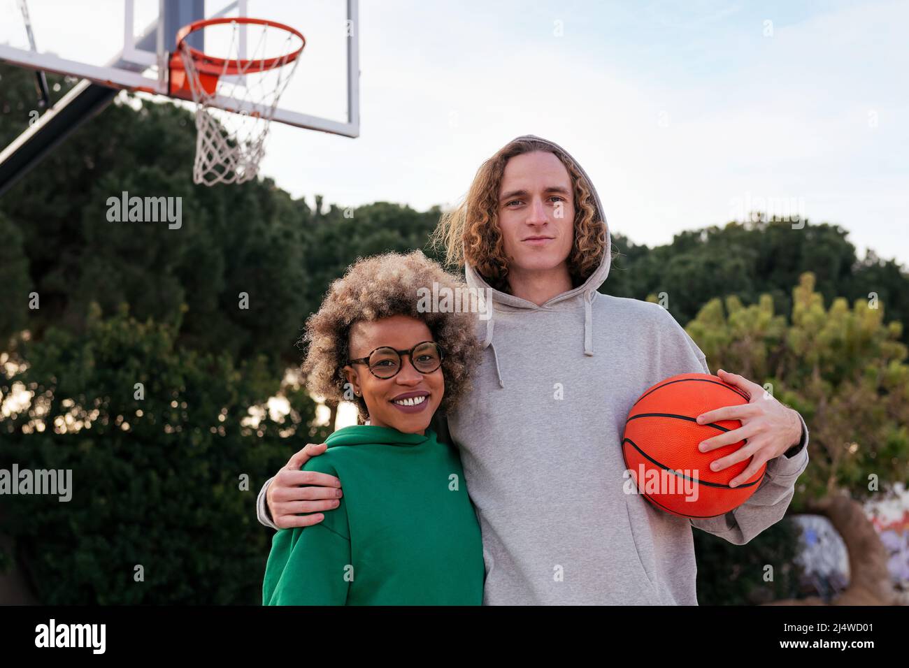 donna latina e uomo caucasico posano sorridendo sul campo di pallacanestro in un parco cittadino, concetto di amicizia e sport urbano Foto Stock
