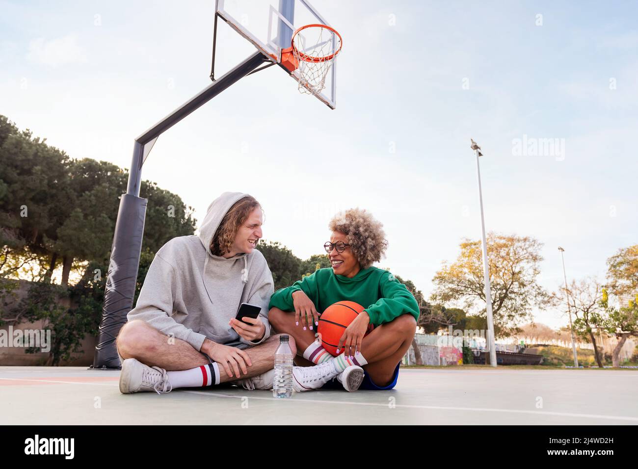 coppia di amici che ridono e si divertono seduti in campo dopo una pratica di basket in un parco cittadino, concetto di amicizia e sport urbano in th Foto Stock