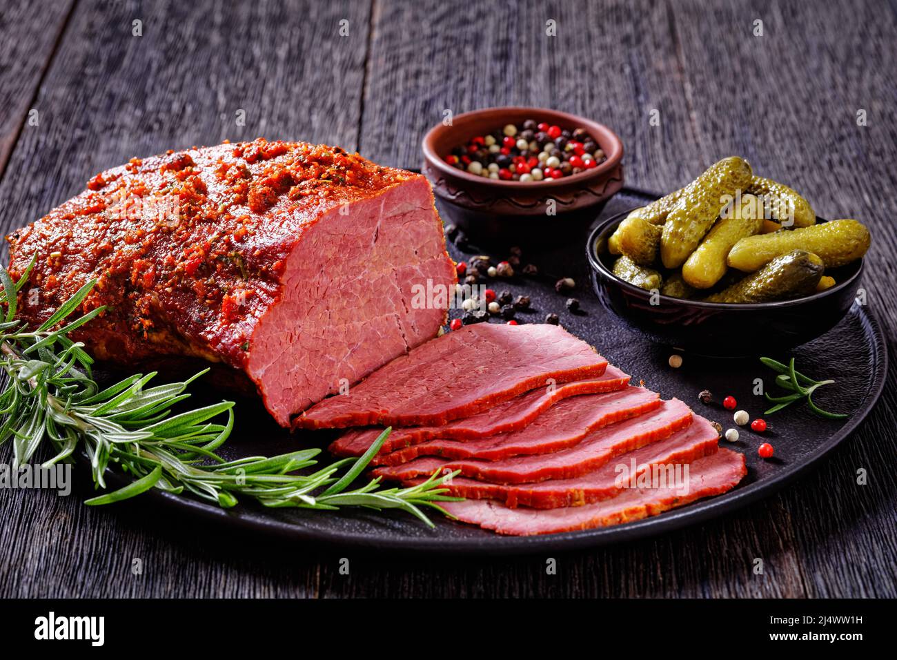 pastrami di manzo arrosto affettati su piatto nero con rosmarino fresco, timo e sottaceti su tavola scura, vista orizzontale dall'alto Foto Stock