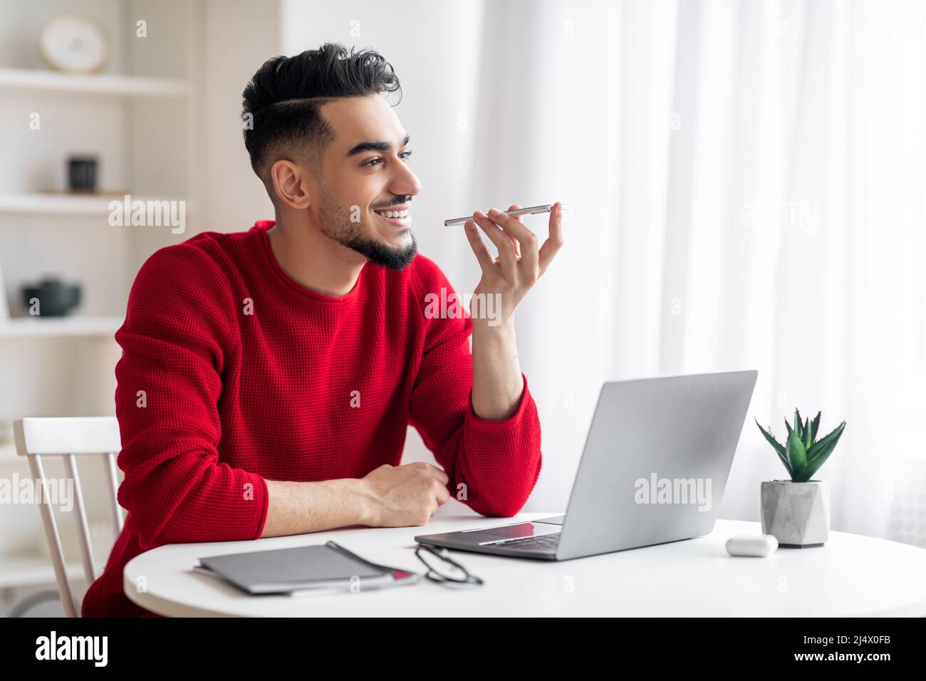 Sorridente ragazzo arabo millenario con barba in abiti rossi parla telefonicamente sul posto di lavoro con un computer portatile Foto Stock