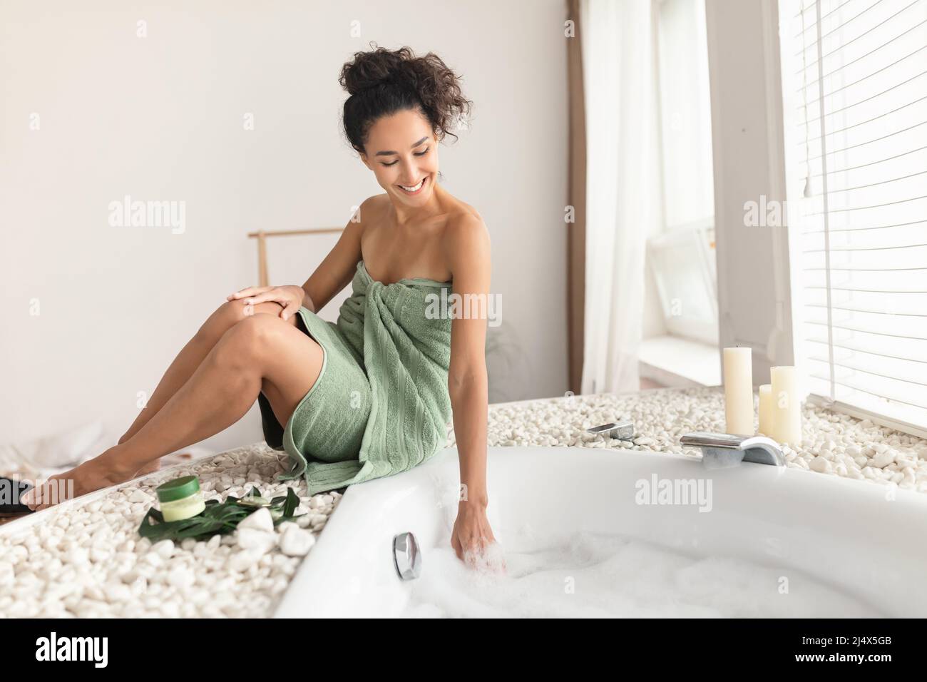 Affascinante giovane donna in asciugamano fare bagno schiumoso, toccare l'acqua, controllare la temperatura, pronto per la procedura termale di casa Foto Stock