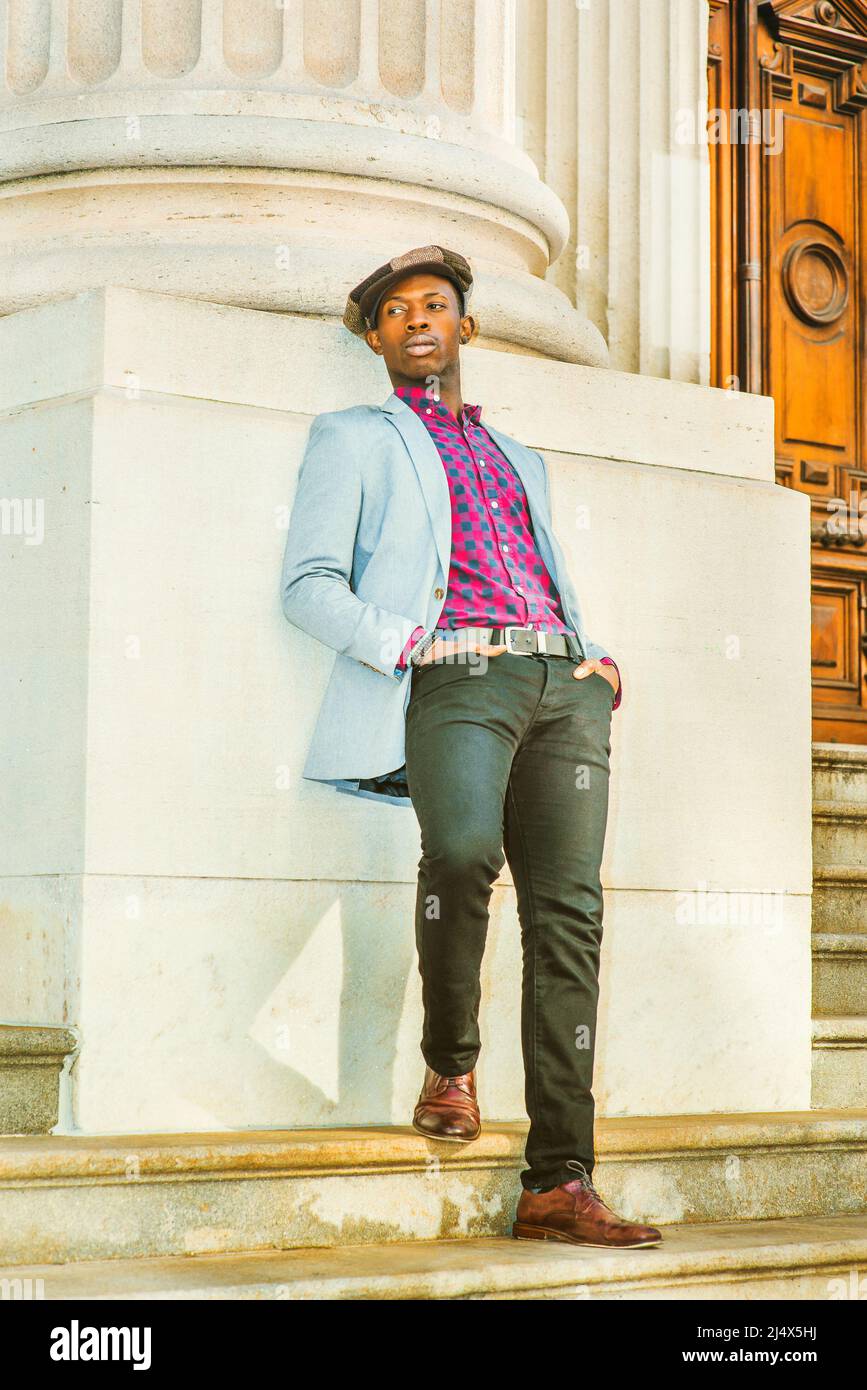 Uomo che si appoggia indietro. Con cappellino da edicola alla moda, vestito in blazer grigio chiaro, rosa a motivi geometrici, maglietta nera, pantaloni neri, scarpe in pelle marrone, Foto Stock