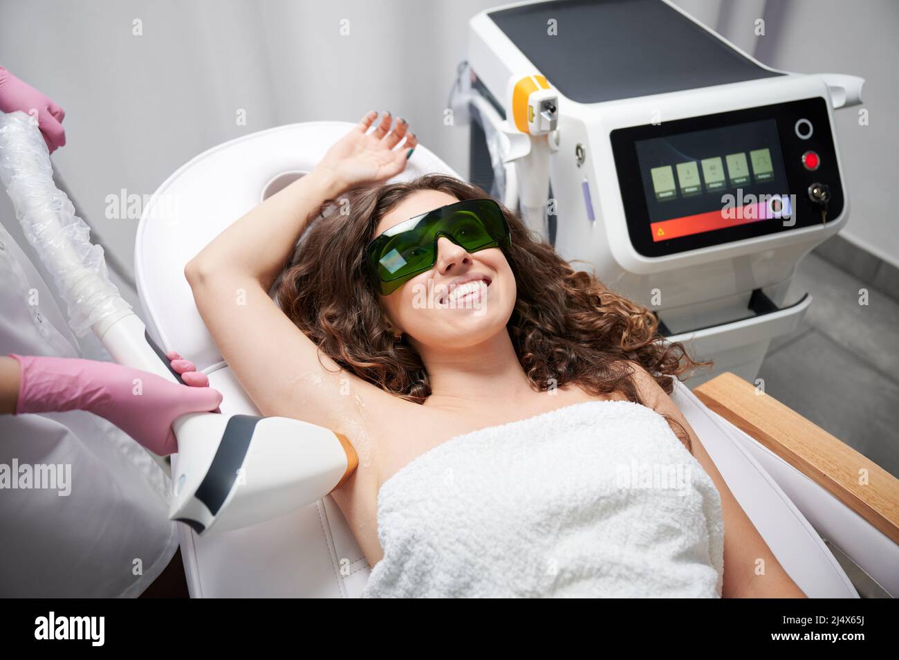 Giovane donna in occhiali di sicurezza sdraiati sul letto da giorno e sorridenti mentre estetista rimuovere i capelli da ascella femmina con dispositivo laser. Cosmetologo che esegue la procedura di rimozione dei capelli laser in salone di bellezza. Foto Stock