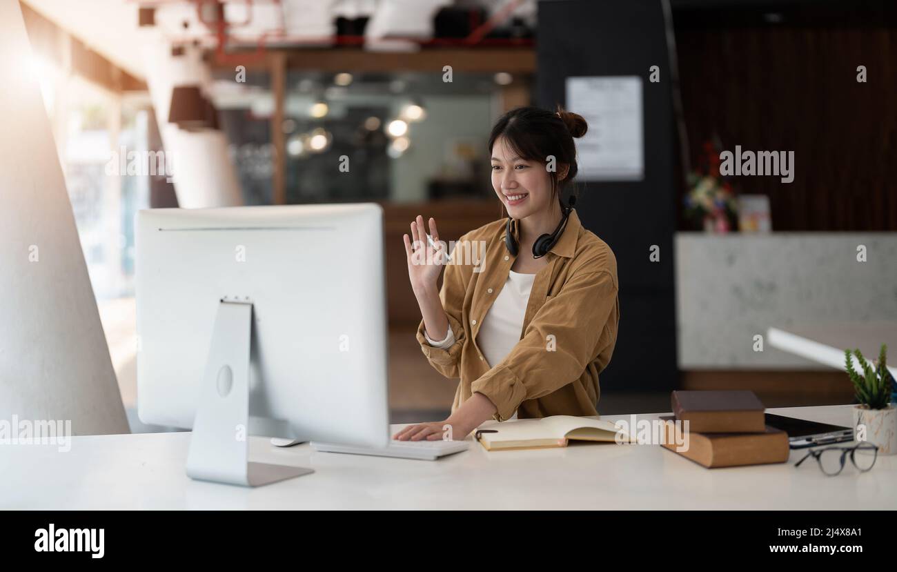 Gioiosa giovane donna asiatica in cuffie wireless che ondeggiano il saluto della bacchetta sullo schermo mentre si siede al tavolo e si ha una video chat con i partner commerciali Foto Stock
