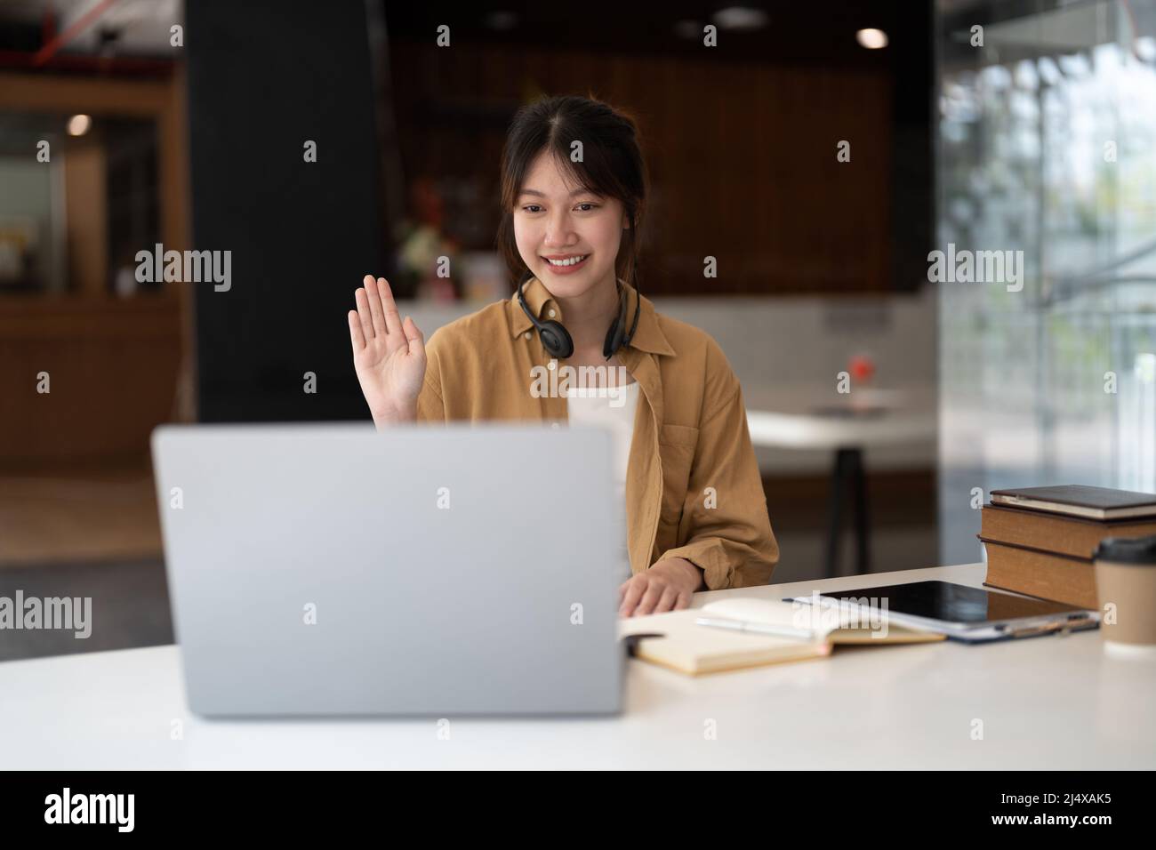 Gioiosa giovane donna asiatica in cuffie wireless che ondeggiano il saluto della bacchetta sullo schermo mentre si siede al tavolo e si ha una video chat con i partner commerciali Foto Stock
