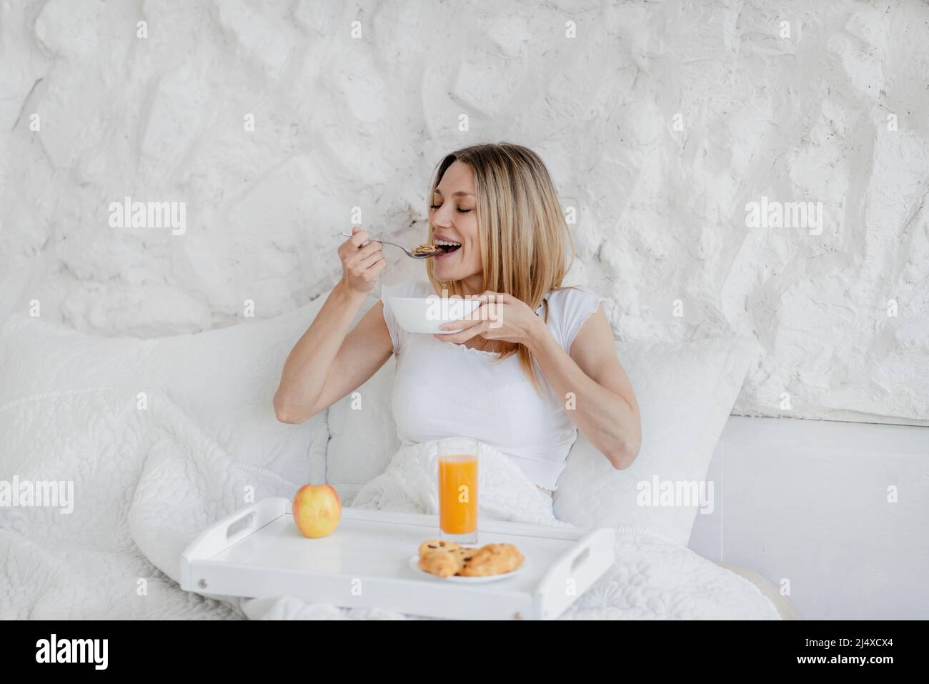 Felice abbastanza affamata giovane donna bionda europea svegliarsi, fare colazione in letto bianco Foto Stock